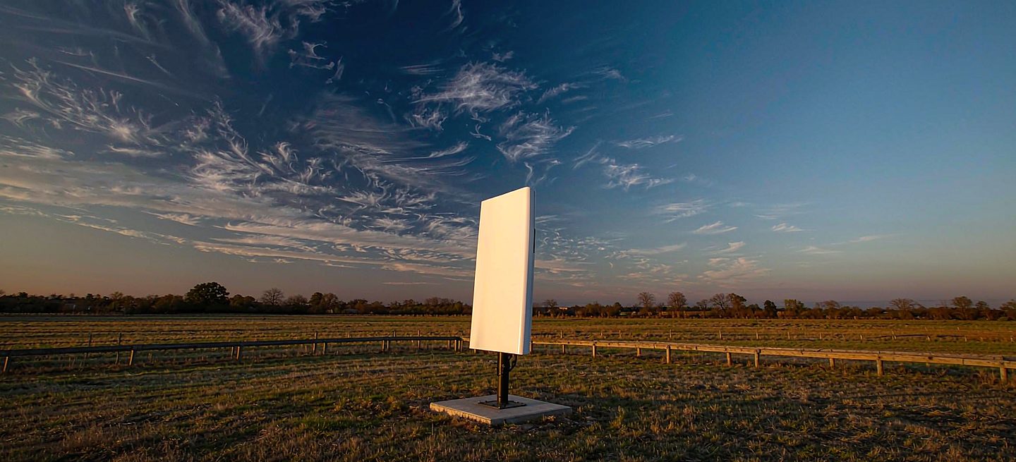 A dish on a raised mount looking over a nearby obstruction toward open sky, with a distant tree line low on the horizon behind it