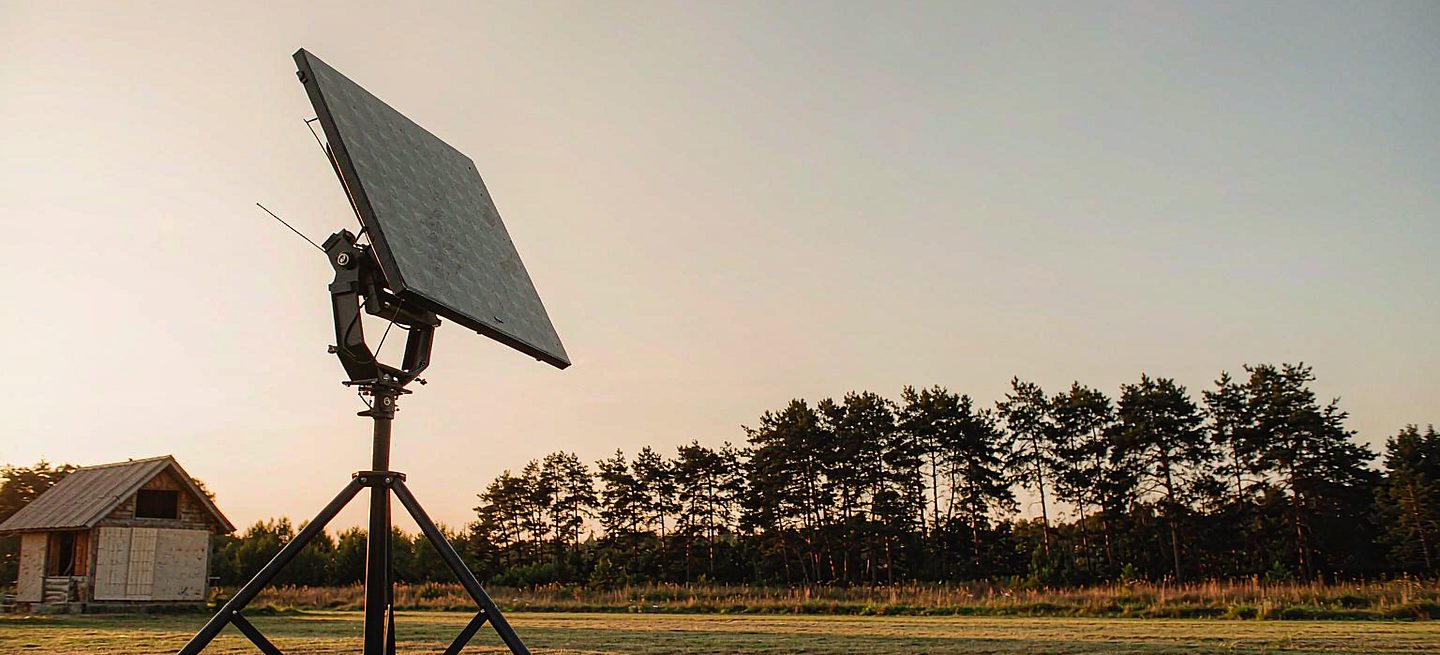 A freestanding Starlink dish on a tripod stand in a back garden at golden hour, aimed at open sky above a pine treeline, with a house left untouched in the background