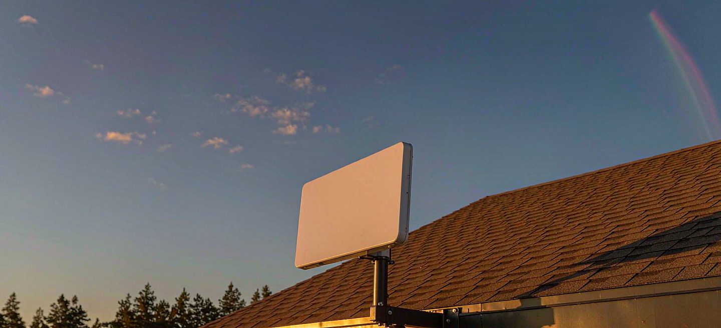 A Starlink-style flat rectangular phased-array panel carried on a black steel arm bracket fixed to the fascia board at the edge of a roof, aimed at open sky above a pine treeline, with the roof covering untouched